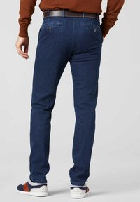Homme portant un pantalon bleu foncé ajusté, une ceinture marron, un pull couleur rouille et des baskets blanches avec des accents bleu marine et marron, debout de dos.