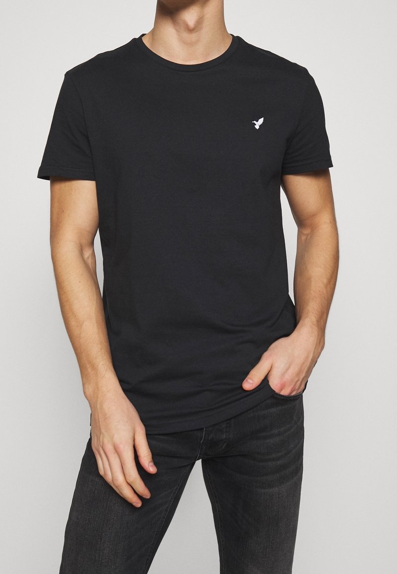 Homme portant un t-shirt noir à manches courtes avec un petit logo d'oiseau blanc sur la poitrine gauche et un jean noir, main dans la poche.