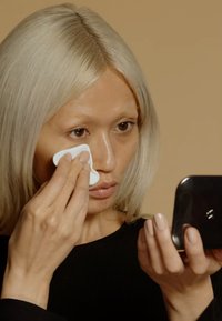 En person med rakt, ljust blont hår applicerar produkt med en vit svamp, med en svart handhållen spegel, mot en dämpad beige bakgrund.