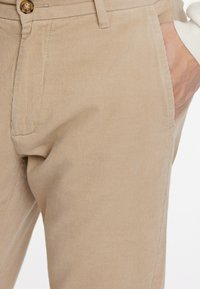 Beige corduroy pants featuring a textured surface, slim fit, subtle vertical stripes, and a front pocket with a button closure.