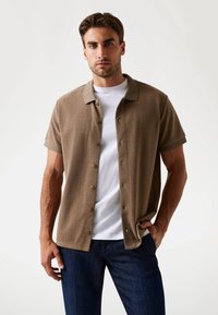 Chemise beige à manches courtes avec un motif en chevrons, associée à un t-shirt blanc et un pantalon bleu marine. Boutons visibles à l'avant de la chemise.
