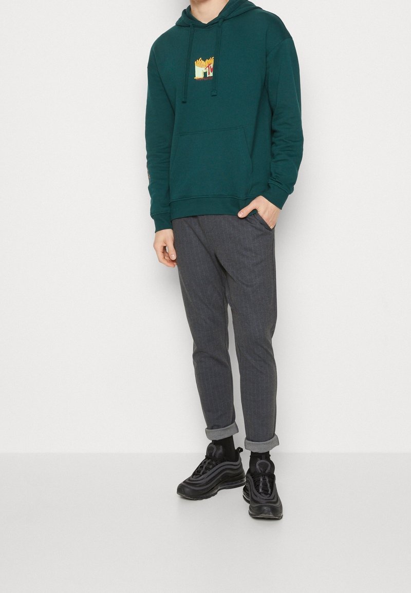 Sweat à capuche bleu sarcelle avec un motif de frites, associé à un pantalon jogger à carreaux gris, retroussé aux chevilles, et des baskets noires avec des semelles distinctives.