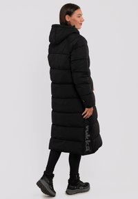 Abrigo de plumas negro con capucha, que presenta un silueta larga y rectangular, textura acolchada y un acento de logo a lo largo del lado. Llevado con leggings negros y botas.