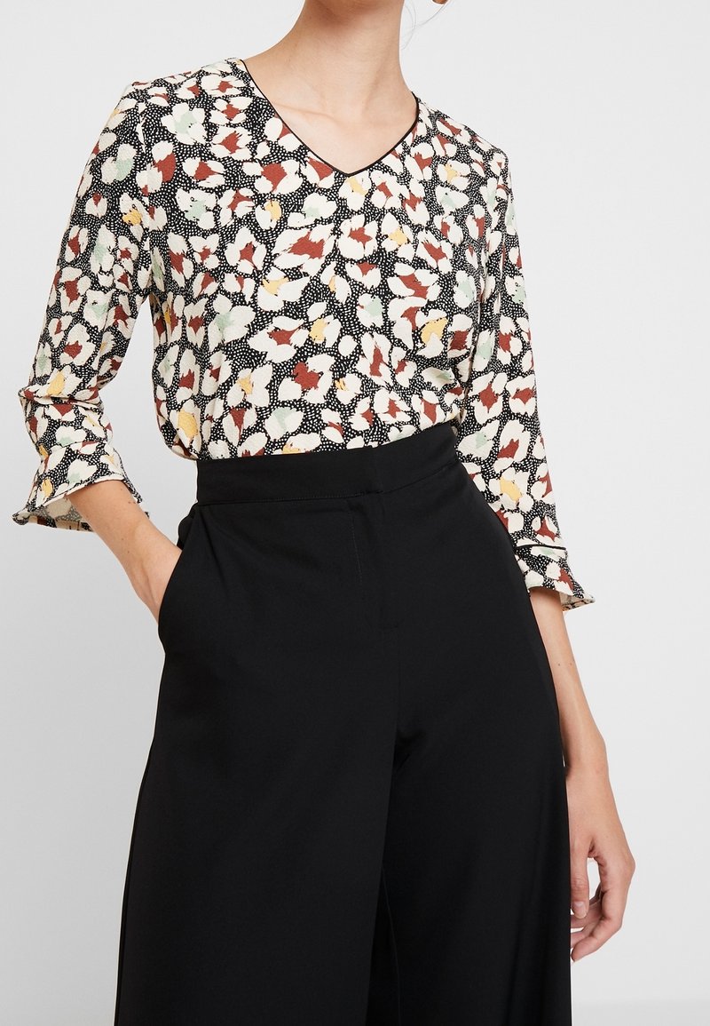 Femme portant un chemisier à col en V à motif floral avec des manches trois-quarts, rentré dans un pantalon noir taille haute à jambes larges.
