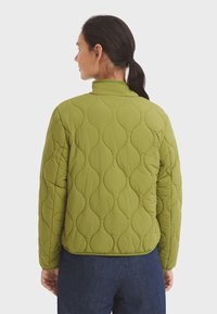 Gequilted groene jas met een hoge kraag, gekenmerkt door een textuur met golvende patronen en elastische manchetten. Draagt over donkere denim broek.