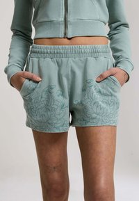 Femme portant un short taille élastique bleu-vert clair avec imprimé floral et un sweat à capuche court zippé assorti, mains dans les poches, et un tatouage de papillon sur la cuisse.