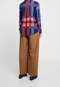 Hemn žíňový s dlouhým rukávem s barevným kostkovaným vzorem v červené, růžové a modré, kombinovaný s širokými hnědými kalhotami a černými botami.