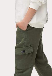 Pantaloni cargo verde oliva con una tasca laterale, tessuto testurizzato, dettagli cuciti e un'etichetta sulla tasca. Indossati con una felpa bianca.