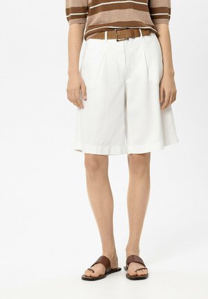 Person wearing white knee-length pleated shorts, brown striped top, brown woven belt, and brown flat sandals standing on white background.