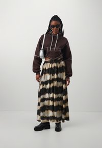 Bruine cropped hoodie met witte trekkoorden, gecombineerd met een lange geplooide rok met een tie-dye patroon in zwart, crème en bruin, en zwarte laarzen.