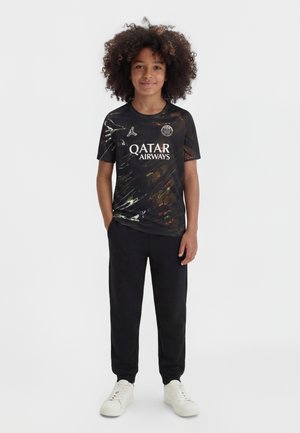Dieťa s kučeravými vlasmi stojí, oblečené v čiernom športovom drese Qatar Airways, čiernych nohaviciach a bielych teniskách na jednoduchom pozadí.