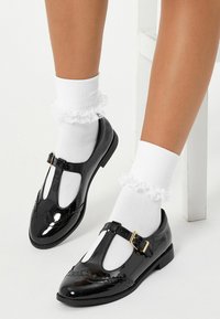 Svarta patenterade läderskor med T-rem och en liten metallspänne, i kombination med vita strumpor med volangkant.