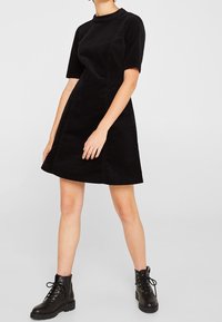 Schwarzes Cordkleid mit kurzen Ärmeln, Rundhalsausschnitt und A-Linien-Silhouette, kombiniert mit schwarzen Schnür-Stiefeletten.
