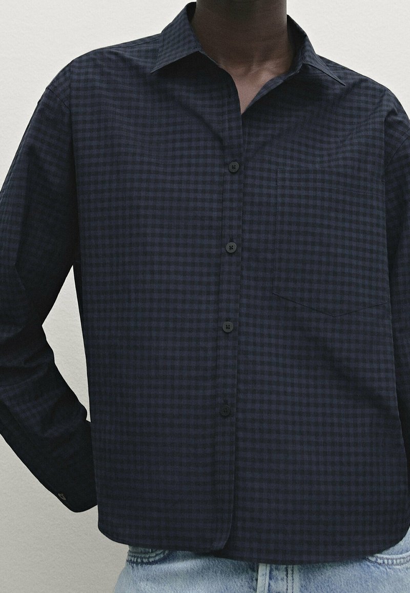 Homme portant une chemise à carreaux bleu foncé et noir avec boutons et poche avant, ainsi qu'un jean en denim bleu clair.