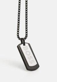 Svart metall hundetags anheng med sølvdetaljer som viser ordet "BOSS." Festet til en kort, svart kjede med lenkedesign.