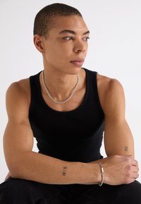 Top cropped preto com nervuras, colar de corrente prateada e pulseira texturizada prateada. O modelo tem cabelo curto e tatuagens visíveis nos braços. Fundo neutro.