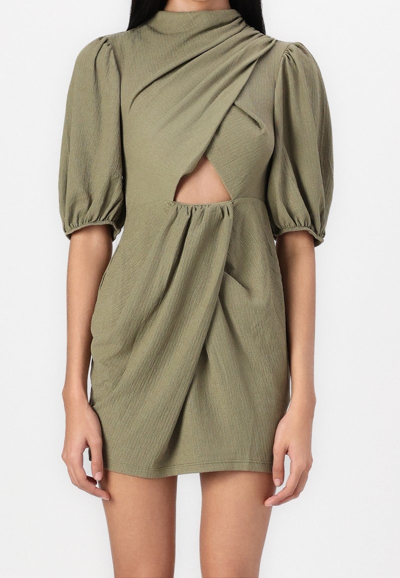Femme portant une mini-robe vert olive avec des manches bouffantes, un col montant et une découpe à l'avant ornée de tissu drapé.