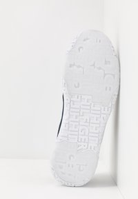 Hvid sneakersålers med tekstureret gummi, der har prægede mærkenavne og geometriske mønstre. Mørkt stof på overdelen er synligt i kanten.
