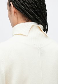 Πουλόβερ σε κρεμ απόχρωση με ριμπ γιακά, που παρουσιάζει απαλή υφή και διακριτική ραφή στο πίσω μέρος.