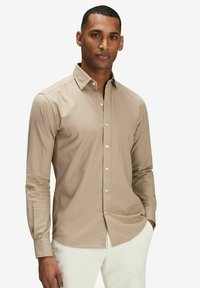 Ljusbrun långärmad skjorta med krage, med en knappfront och en slät yta. Bärs med vita byxor.