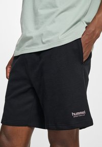 Shorts de sport noirs en mélange de coton, avec des poches latérales et un accent de logo rose sur l'ourlet. Association avec un haut vert clair.
