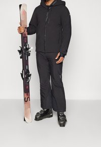 Osoba v černé lyžařské bundě, kalhotách a botách, držící pár růžových a černých lyží vztyčených proti jednolitému pozadí.