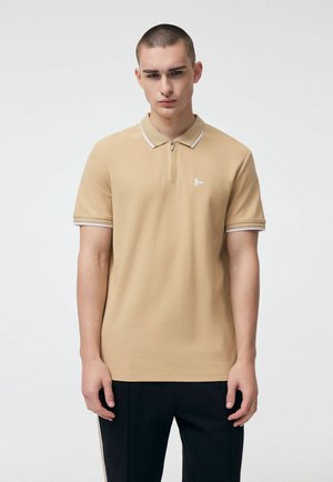 Młody mężczyzna w beżowej koszulce polo z krótkim rękawem na zamek oraz czarnych spodniach, stojący na tle gładkiej białej ściany.