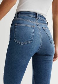 Mujer que lleva jeans ajustados azul de cintura alta con bolsillos traseros y una camiseta de tirantes blanca acanalada, mostrada desde la cintura hasta la mitad del muslo.