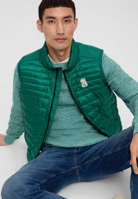 Grüne gesteppte Weste mit hohem Kragen, Reißverschluss und kleinem Logoschild. Über einem strukturierten hellblauen Pullover und blauen Jeans getragen.