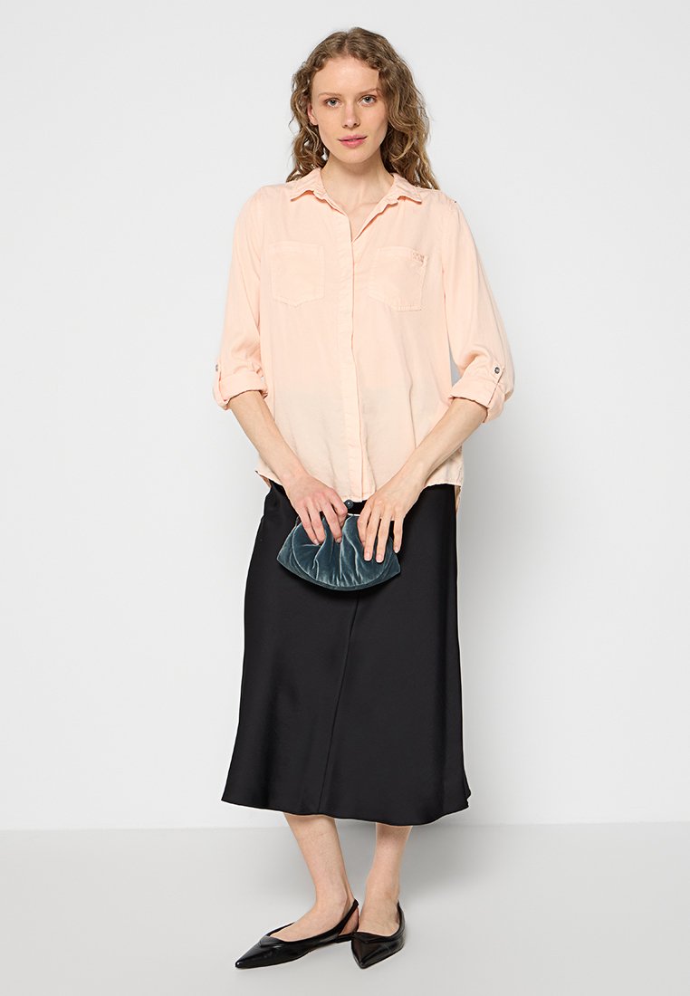 Camisa de manga larga rosa claro con dos bolsillos en el pecho, combinada con una falda midi de satén negra. Sosteniendo un bolso de terciopelo en tono azul verdoso, con zapatos planos negros.