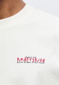 Κοντινή λήψη λευκού T-shirt με κόκκινο και μαύρο κείμενο που γράφει "5 Αυγούστου Kräftskiva hosted by Marc O’Polo Denim" στο στήθος.