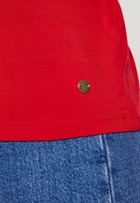 Camicia rossa a maniche corte in morbido tessuto, con un piccolo badge in bronzo con la scritta "ESPRIT". La camicia è abbinata a un denim blu texturizzato.