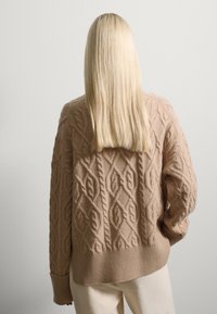 Suéter de punto beige con patrones de trenzado en relieve, dobladillo de canalé y hombros caídos. La textura parece suave y gruesa.