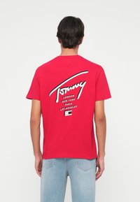 Červené bavlnené tričko s veľkým bielym grafickým logom na chrbte. Výrazný text obsahuje "Tommy" a názvy miest v čiernej farbe. Voľný strih.