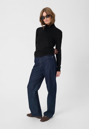 Crna majica s dugim rukavima i lažnim turtleneck ovratom u kombinaciji s tamnoplavim širokim trapericama. Model nosi crne naočale i smeđe mekane cipele.