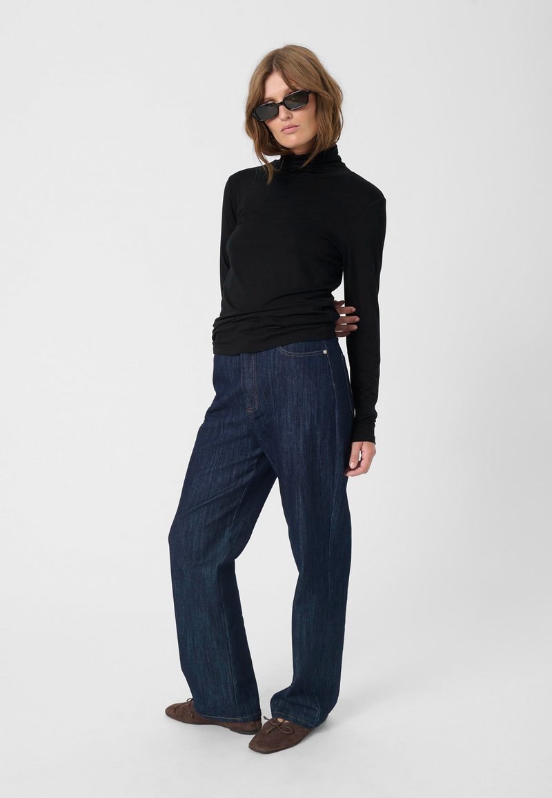 Black long-sleeve mock turtleneck top paired with dark blue wide-leg denim jeans. Model wears black sunglasses and brown soft shoes.