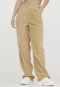 Pantalon en velours côtelé beige à jambes larges, doté d'une taille haute, de deux poches avant et d'une texture lisse. Associé à des baskets blanches à plateforme.