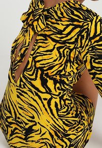 Šaty se zebrovým vzorem v žluté a černé barvě, s halter krkem, výřezy na bocích a mašlí u výstřihu. Hladká, elastická látka.