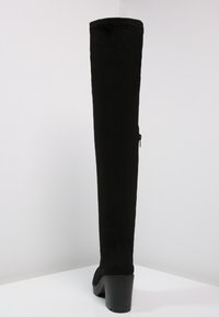 Botín negro de muslo alto, hecho de material suave, con un tacón grueso y un detalle de cremallera lateral, con una textura suave en toda su superficie.