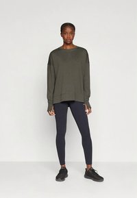 Volné mikina olivově zelené barvy s spadlými rameny, doplněná černými legínami. Oba oděvy mají hladkou texturu.