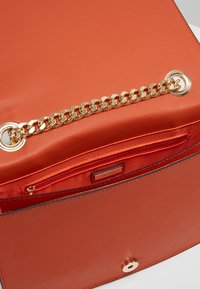 Prostokątna pomarańczowa skórzana torebka z złotym łańcuszkiem i wewnętrzną kieszenią na zamek. Gładka faktura i czyste krawędzie.