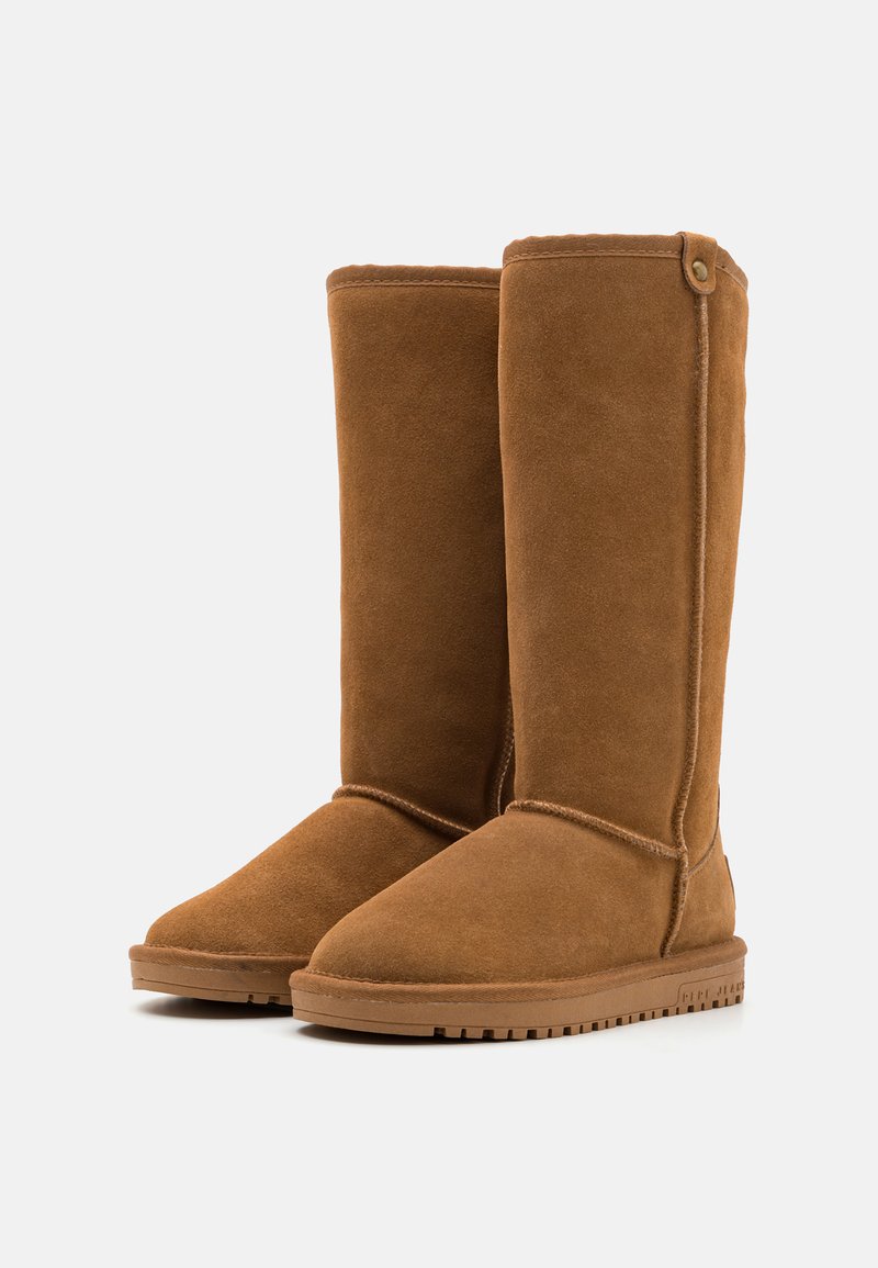 Bottes en daim marron jusqu'au genou avec une semelle plate beige, un bout arrondi et des coutures surpiquées. Présente un bouton décoratif sur le côté.