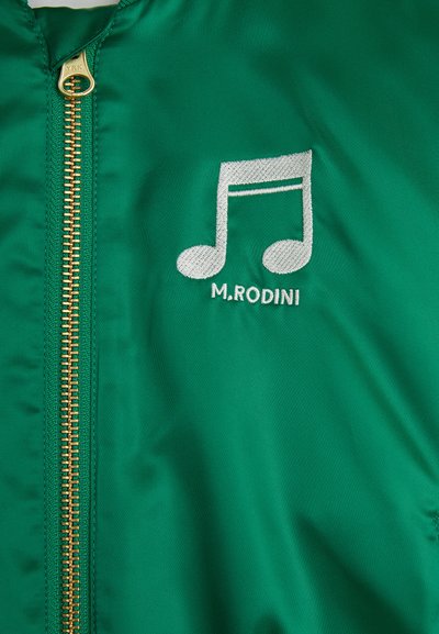 Zelena bomberska jakna z zlatim zadrgo, ki na levi strani prsi vsebuje belo vezeno glasbeno noto in besedilo "M.RODIN".