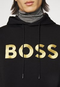 Felpa nera con cappuccio caratterizzata da grandi lettere ricamate in oro che formano la parola "BOSS". Include un laccio con estremit�à nere. Sotto, un dolcevita a righe bianche e nere.