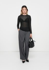 Black sheer long-sleeve top with a v-shaped design, paired with gray wide-leg trousers. Model holds a black textured handbag.