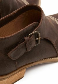 Botas de tobillo de cuero marrón con una superficie texturada, que presentan un detalle de hebilla metálica y una suela de color claro. Se aprecian acentos de costura.