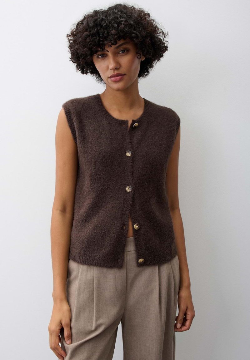 Woman with curly hair wearing a sleeveless brown buttoned vest and beige high-waisted trousers standing against a plain background.