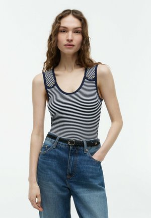 Femme portant un top sans manches rayé bleu marine et blanc avec des boutons dorés sur les bretelles, un jean bleu taille haute et une ceinture noire tressée.