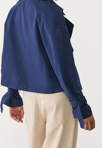 Granatowa, krótka kurtka z gładkiego materiału, z szerokimi mankietami z zapięciem na troczki, noszona z beżowymi spodniami o wysokim stanie.