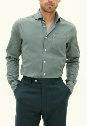 Camicia verde chiaro con bottoni e taschino sul petto, abbinata a pantaloni su misura verde scuro. Tessuto liscio, vestibilità casual e bottoni a contrasto.